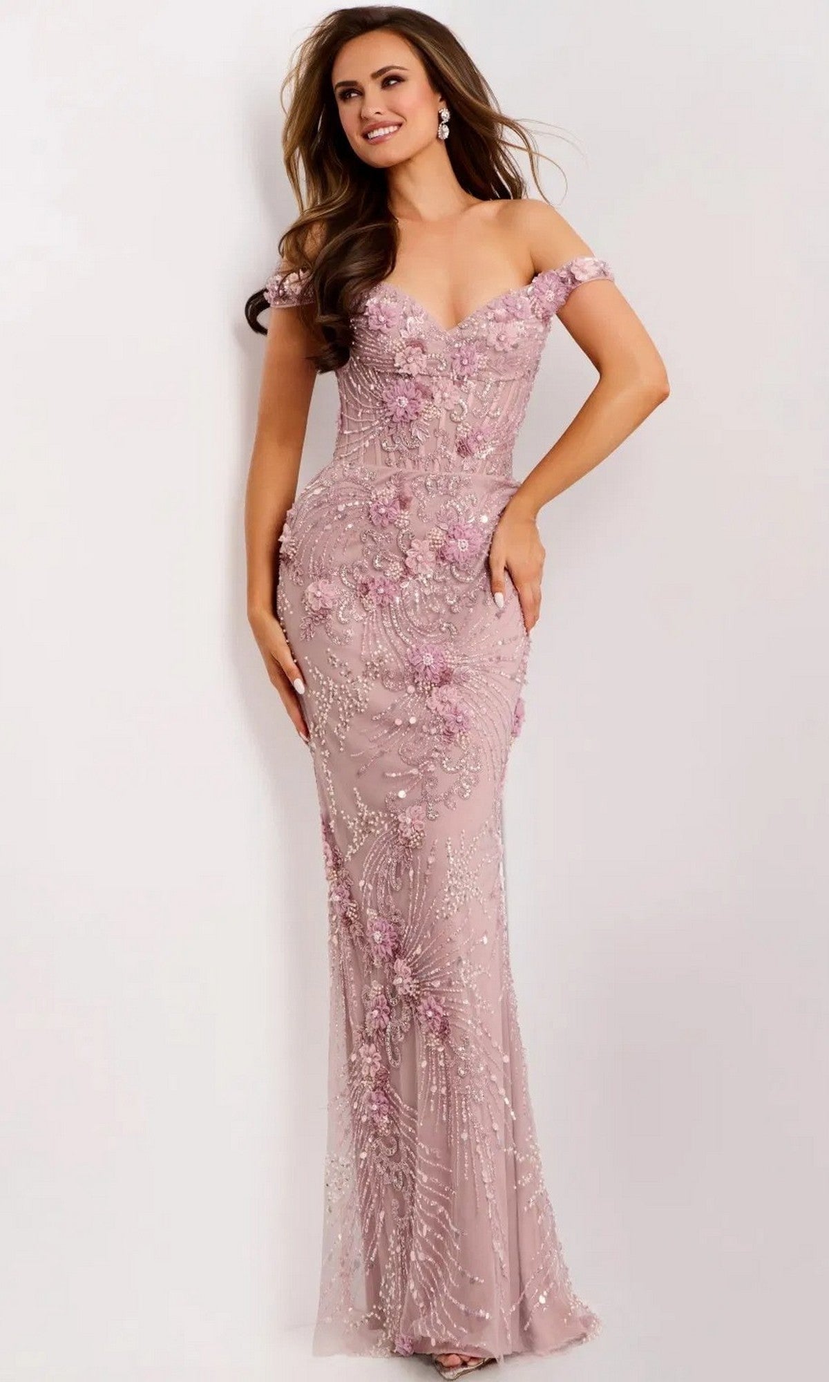 A woman wears the Jovani 44007 Long Prom Dress, a fitted off-the-shoulder pink gown featuring delicate beadwork and 3D floral appliques, as she stands against a plain light background.
